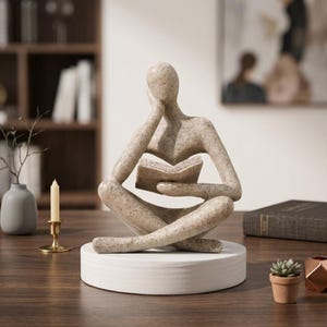 Könnte beinhalten: Eine beige gesprenkelte abstrakte Skulptur einer sitzenden Figur, die ein offenes Buch liest. Die Figur stützt ihren Kopf auf eine Hand und sitzt im Schneidersitz auf einem glatten, runden weißen Sockel mit einer strukturierten Oberfläche.