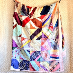 May include: A patchwork quilt constructed from various fabric scraps in a multitude of colours including red, blue, purple, yellow, and pink. The pieces are arranged in a diagonal pattern and joined together with visible zigzag stitching throughout.