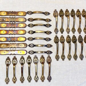 May include: A collection of antique-style brass hardware including seven elongated rectangular backplates with two screw holes each, six arched drawer pulls, and twenty vertical drop-style cabinet handles with ornate mounting plates.