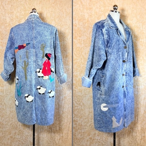May include: A long-sleeved acid-wash denim coat featuring a button-down front and a collar. The back displays a colorful applique scene of a figure in a red coat tending to a flock of white sheep among cacti, while the front shows a moon and wolf.