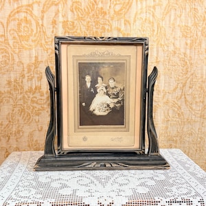 May include: A black swivel picture frame holding a sepia-toned vintage photograph of four people. The frame features curved side supports and a rectangular base. The photograph depicts a group portrait with a central figure in a light-colored dress.