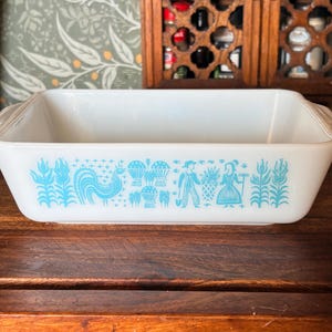 May include: A rectangular white glass baking dish featuring a repeating light blue pattern along the side. The design includes illustrations of a rooster, stalks of corn, wheat sheaves, and a pair of figures standing near a basket of produce.