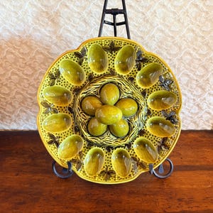 Könnte beinhalten: Eine Vintage-Eier-Servierplatte aus Keramik. Die Platte ist leuchtend gelb mit olivgrünen, eiförmigen Vertiefungen. Die Mitte hat eine Ansammlung von olivgrünen, eiförmigen Vertiefungen. Die Platte steht auf einem schwarzen Metallständer.