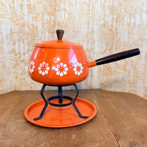 May include: A bright orange fondue pot with a white floral pattern, featuring a matching lid with a brown knob and a long black handle. The pot rests on a black metal stand that is positioned on a circular orange metal base plate.