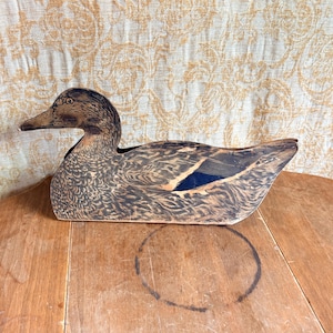 May include: A flat, wooden duck silhouette featuring detailed brown and black feather patterns across the body, a dark head, and a distinct blue patch on the wing. The object is shaped like a swimming mallard and has a smooth, painted finish.