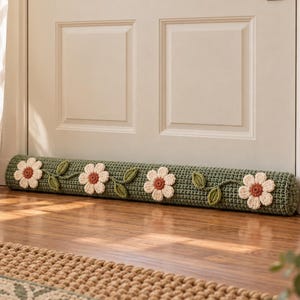 May include: A long, cylindrical door draft stopper featuring a sage green crocheted base decorated with four white flowers with rust-colored centers and green crocheted leaves connected by a vine pattern along the length of the fabric.