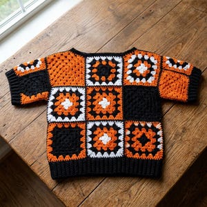 May include: A short-sleeved crochet top constructed from a grid of square motifs in black, bright orange, and white. The garment features a crew neckline and ribbed black trim along the sleeves and bottom hem, showcasing a geometric granny square pattern.