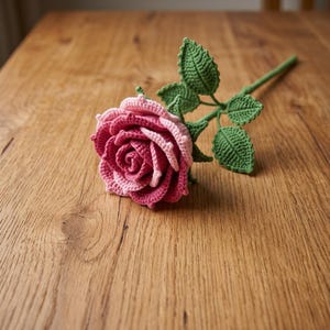 May include: A handcrafted crochet rose featuring layered petals in shades of deep magenta and soft pink. The flower is attached to a long green stem with three textured, pointed green leaves extending from the stalk, showcasing intricate yarn work.