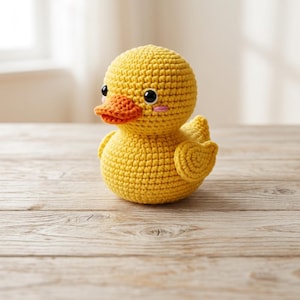 May include: A small, round, yellow crocheted duck toy with a textured yarn surface. The duck features a bright orange beak, two glossy black circular eyes, and small pink embroidered blush marks on its cheeks. It has a rounded body and wings.