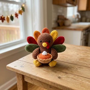 May include: A crocheted turkey figurine featuring a brown body, a yellow beak, and a red wattle. The turkey has a fan of colorful feathers in red, yellow, and green, and is holding a small orange pumpkin pie topped with a white dollop of cream.