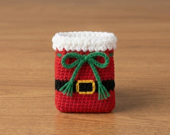 Puede incluir: Una pequeña bolsa cilíndrica de ganchillo con cuerpo rojo y un borde texturizado blanco en la parte superior. El diseño incluye una banda horizontal negra con el contorno de una hebilla rectangular amarilla y un lazo verde de ganchillo.
