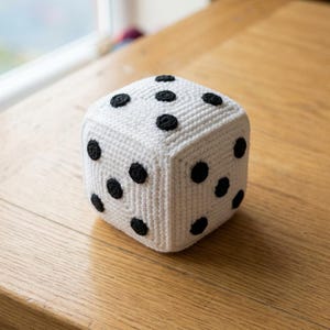Pode incluir: Um cubo de crochê branco em forma de dado de seis lados com pontos circulares pretos costurados nas faces. A textura do fio é visível em toda a superfície do objeto, que apresenta cantos arredondados e uma aparência macia e artesanal.
