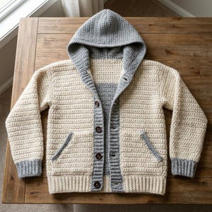 May include: A cream-colored crochet hooded cardigan featuring long sleeves with grey cuffs, a grey hood, and a grey button-down placket. The garment includes two front pockets with grey trim and four dark brown buttons down the center front.