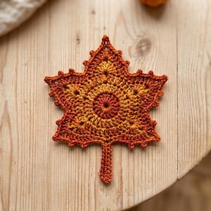 May include: A crochet maple leaf coaster featuring a gradient of warm autumn colors including deep orange, burnt red, and golden yellow. The piece has a textured, circular center with pointed, jagged edges and a small stem at the bottom.