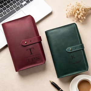 May include: Two leather journals, one burgundy and one dark green, each featuring a strap closure with a brass stud. The burgundy journal is engraved with T and TPN, while the dark green journal is engraved with K and Vincent. Both have visible stitching.