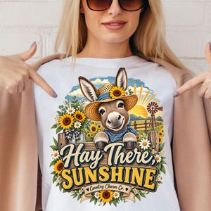 Könnte beinhalten: Ein weißes T-Shirt mit einer Grafik eines Esels mit Strohhut, umgeben von Sonnenblumen und einer Windmühle. Der Text lautet Hay There SUNSHINE und Country Charm Co. Das Design zeigt zudem einen Holzzaun und eine leuchtend gelbe Sonne.