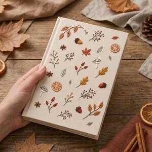 Pode incluir: Um livro de capa dura retangular com uma capa de tecido na cor creme, apresentando motivos bordados com temática de outono, incluindo fatias de citrinos laranja, bolotas castanhas, folhas de ácer vermelhas, cogumelos verdes e anis estrelado.