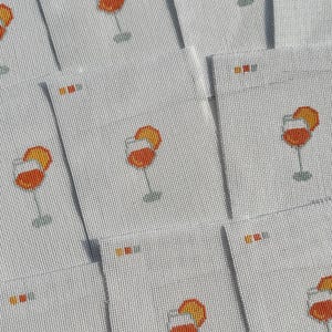 May include: A collection of white needlepoint canvases featuring a repeating pattern of an orange cocktail in a stemmed glass with a citrus slice garnish. Each canvas includes a small color palette guide printed in the upper left corner.