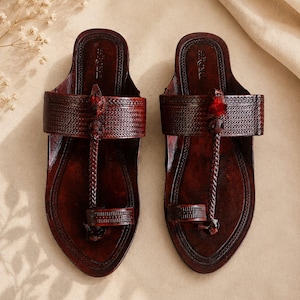 May include: A pair of dark brown leather sandals featuring an intricate embossed pattern on the wide strap and toe ring. The design includes a central braided leather cord connecting the toe loop to the strap, accented with a small red tassel detail.