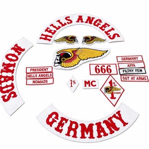 Op de afbeelding: Een verzameling geborduurde patches met de tekst HELLS ANGELS, NOMADS en GERMANY in rood op witte stof. Bevat een gevleugeld schedel-embleem, een 1% ruitvormige patch en rechthoekige patches met tekst zoals PRESIDENT, 666 en SGT AT ARMS.
