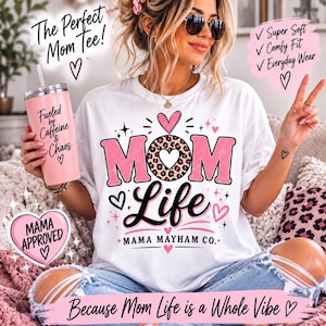 以下が含まれることがあります： 「MOM Life」と「MAMA MAYHAM CO.」の文字がピンクと黒で描かれた白いTシャツ。ヒョウ柄とハートのアクセントが特徴です。「Fueled by Caffeine & Chaos」と書かれたピンクのタンブラーを手に持っています。