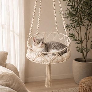 May include: A cream-colored macrame hanging cat bed featuring a circular base with a woven lattice pattern, suspended by three twisted rope cords. A soft, matching circular cushion sits inside the frame, providing a resting spot for a cat.