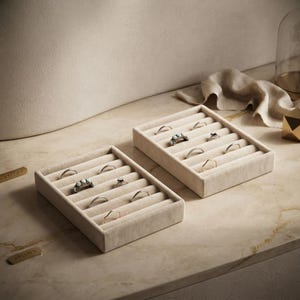 May include: Two rectangular beige velvet ring display trays with multiple slots. The trays are filled with various metal rings, including some with white stones. The trays are positioned on a smooth, light-colored surface with a soft fabric nearby.