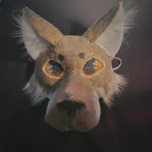 Op de afbeelding: Een beige en goudkleurig dierenmasker met puntige oren met witte imitatiebontrand, een zwarte neus en gaas-oogbedekkingen met oranje en witte details. Het masker heeft een gestructureerd oppervlak en een elastische band voor bevestiging.