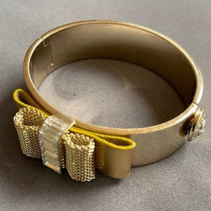 May include: A gold bangle bracelet with a yellow bow accent. The bow has a mesh design and clear rhinestones.