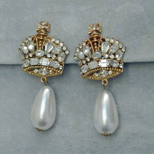 May include: Gold-coloured crown earrings, adorned with clear crystal stones and a large, pear-shaped faux pearl drop. The earrings have a vintage style and are set against a soft grey backdrop.
