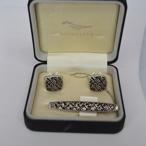 May include: A black box with a white interior containing a silver tie clip and cufflinks. The cufflinks and tie clip have a black and silver geometric design. The box has the word "Kalisvinka" printed on the lid.