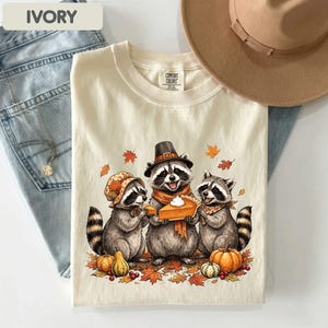 May include: An ivory t-shirt featuring a graphic of three raccoons wearing autumn accessories and holding a slice of pumpkin pie. The central raccoon wears a pilgrim hat and scarf, while the others wear a floral bonnet and a patterned neckerchief.
