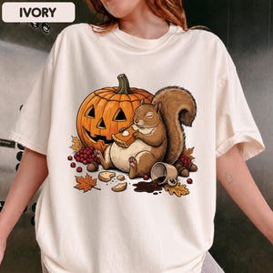 May include: An ivory t-shirt featuring a graphic of a brown squirrel sitting next to a carved orange jack-o'-lantern. The squirrel is eating a slice of pumpkin pie, with a spilled mug of coffee, scattered autumn leaves, berries, and acorns nearby.