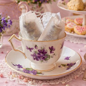 May include: A delicate teacup and saucer set with a floral design, filled with tea bags. The cup and saucer are white with purple flower accents and gold trim. A small bouquet of purple flowers and a tiered plate of cookies complete the scene.