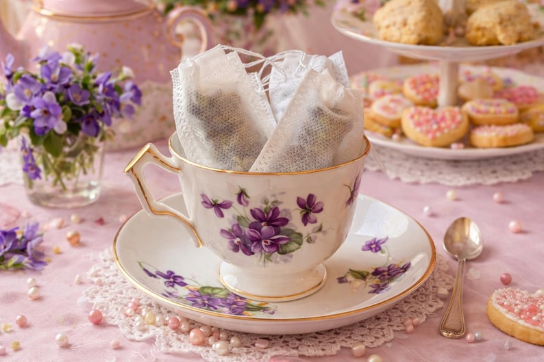May include: A delicate teacup and saucer set with a floral design, filled with tea bags. The cup and saucer are white with purple flower accents and gold trim. A small bouquet of purple flowers and a tiered plate of cookies complete the scene.
