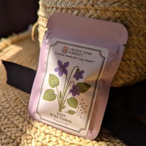 May include: A light purple seed packet featuring a botanical illustration of wild violets. The packet has the text "VELVET VINE MARKET" and "Curious Seeds for Cozy Souls". The packet also has the text "SEEDS Wild Violets (Viola sororia)". The packet is resting on a woven straw surface.