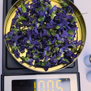 May include: A gold-colored scale pan filled with dried blue and green flowers. The digital display shows a weight of 1.005 grams. The scale is black with a white surface.