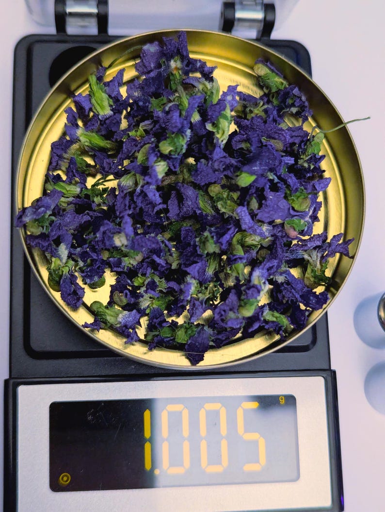 May include: A gold-colored scale pan filled with dried blue and green flowers. The digital display shows a weight of 1.005 grams. The scale is black with a white surface.