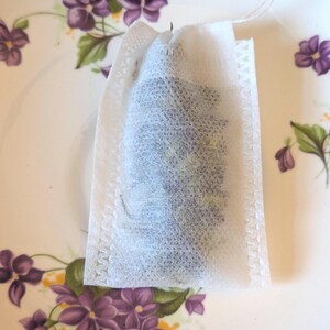 May include: A white tea bag filled with loose tea sits on a vintage plate. The plate has a cream-colored background and is decorated with purple floral patterns. The tea bag is made of a mesh-like material.
