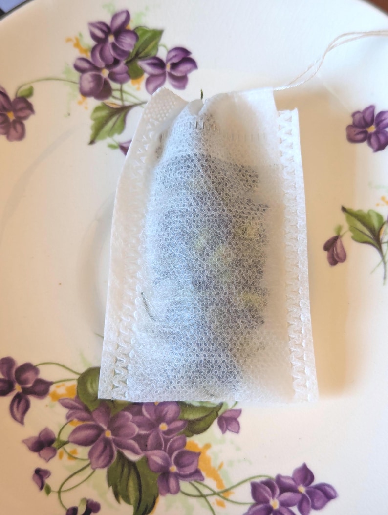 May include: A white tea bag filled with loose tea sits on a vintage plate. The plate has a cream-colored background and is decorated with purple floral patterns. The tea bag is made of a mesh-like material.