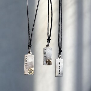 以下が含まれることがあります： 黒いコードに吊るされた3つの長方形のシルバーペンダント。2つのペンダントには小さな金色の足跡と日付「24.10.23」「24.11.15」が刻印されています。3つ目のペンダントには「MY PUPPY」という文字が刻印されています。各ピースは質感のある仕上げです。