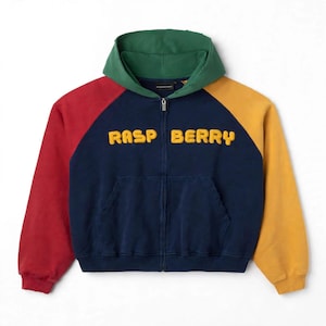 May include: A color-blocked zip-up hoodie featuring a navy blue body, a green hood, a red left sleeve, and a yellow right sleeve. The word RASP BERRY is embroidered in yellow, puffy, rounded letters across the chest. It includes a front kangaroo pocket.