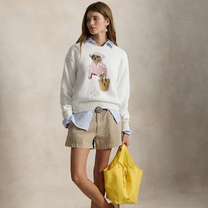 May include: A white long-sleeved sweater featuring a graphic of a bear wearing a striped shirt and a headscarf, layered over a light blue collared shirt. The outfit includes khaki shorts with a decorative belt buckle and a bright yellow tote bag.
