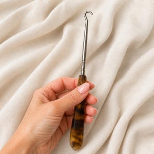 Puede incluir: Un gancho para botones con una punta metálica plateada, delgada y curva, unida a un mango liso y cónico con un patrón de carey en tonos marrones y negros. Esta herramienta está diseñada para abrochar botones en prendas de vestir.