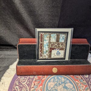 May include: A square card featuring a collage of patterned paper segments in shades of blue, brown, and white, accented with glitter, and a small illustration of a blue bird on a branch, displayed in a wooden stand with a black velvet interior.