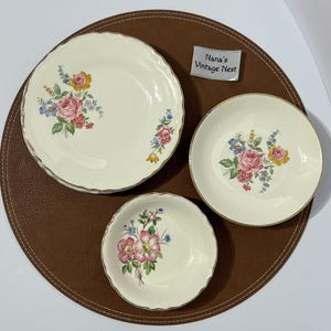 May include: A set of three cream-colored ceramic dishes with scalloped edges and floral patterns featuring pink roses and blue accents. A small white tag with the text Nana's Vintage Nest is included. The dishes have gold-toned rims.