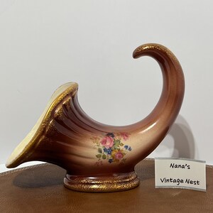 May include: A brown cornucopia-shaped ceramic vase with a cream-colored interior and gold-toned trim along the base and rim. The exterior features a floral decal with pink roses and blue flowers. A small white card displays the text Nana's Vintage Nest.