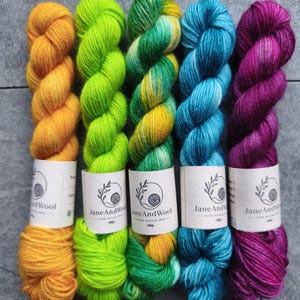 Könnte beinhalten: Fünf nebeneinander angeordnete Garnstränge, jeder mit einem weißen Papieretikett mit dem Text JaneAndWool versehen. Die Farben von links nach rechts sind sattes Goldgelb, helles Limettengrün, meliertes Grün-Gelb, sattes Blau und tiefes Magenta.