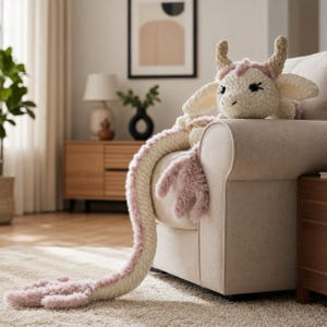 May include: A long, plush, crocheted dragon toy featuring a cream-colored body with soft, dusty pink accents along its spine, tail, and head. The creature has two horns, large dark eyes, and a long, winding tail that extends down to the floor.