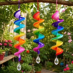 May include: Three hanging spiral decorations made of crocheted yarn in a rainbow gradient of colors. Each spiral features a clear, faceted teardrop-shaped crystal pendant at the bottom and is suspended by a matching yarn loop at the top.